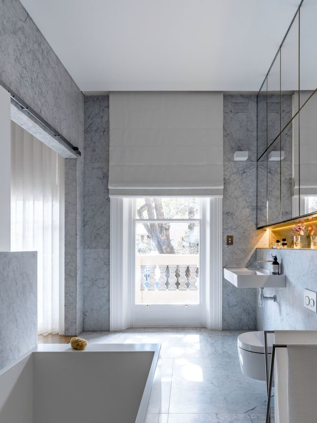 Bright, luxurious contemporary bathroom clad in pale grey-white marble, with a freestanding bathtub in the foreground, wall-mounted toilet, floating vanity, large mirrored cabinetry, and a tall window with a roller blind. Natural light enters through sheer curtains and the window, creating a refined spa-like atmosphere.