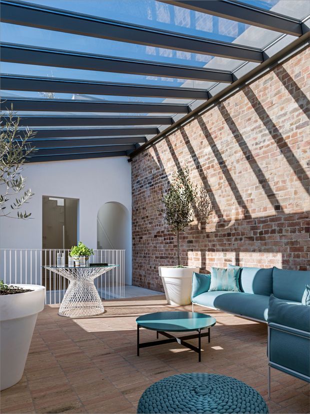 A bright enclosed courtyard or terrace with a transparent glazed roof supported by dark metal beams. The space combines an exposed red brick wall with white rendered walls, terracotta-toned tiled paving, white planters, and a contemporary teal outdoor sofa, glass-topped coffee table, and woven side table. Strong linear shadows from the roof structure create a graphic pattern across the brick wall and floor. A small arched opening and railing are visible at the rear.