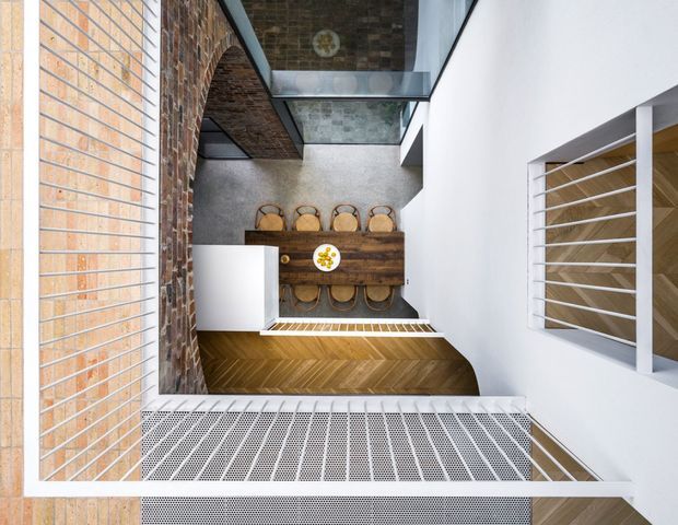 A top-down view of a contemporary dining area within a narrow, multi-level interior courtyard or void. A long dark timber dining table with multiple woven dining chairs is positioned beside a white kitchen island or built-in counter. Exposed red brick walls, white plaster walls, a glazed roof or skylight, timber herringbone flooring, and perforated metal grilles create a strong mix of industrial and refined modern detailing.