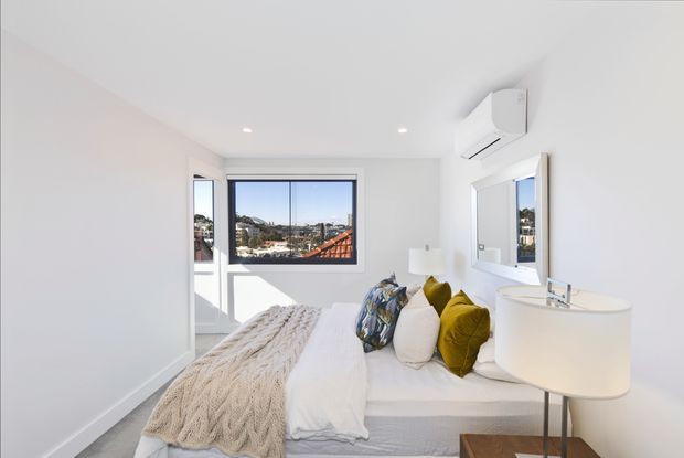 Bright contemporary bedroom with a neatly made bed, white bedding, textured beige throw, and mustard and patterned accent cushions. The room has white walls and ceiling, recessed downlights, a wall-mounted air-conditioning unit, bedside table and lamp, and a large window overlooking a dense urban residential setting.