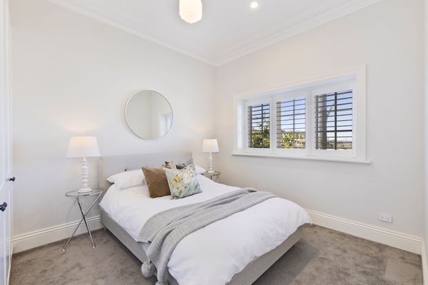 Bright, compact bedroom with a neatly made bed, soft grey throw, neutral cushions, mirrored bedside tables with lamps, a round wall mirror, white plantation-style shutters, carpeted flooring, and white decorative ceiling and wall trim.