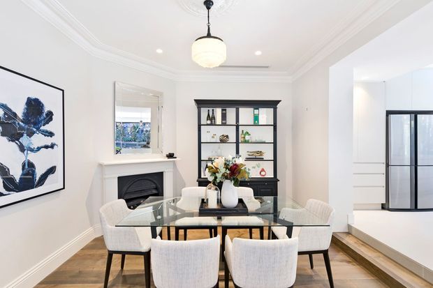 Bright, refined dining room with a rectangular glass dining table and upholstered chairs, a decorative fireplace with mantel, built-in dark shelving, framed artwork, pendant lighting, and detailed ceiling cornices. An open kitchen is visible through the wide opening at right.