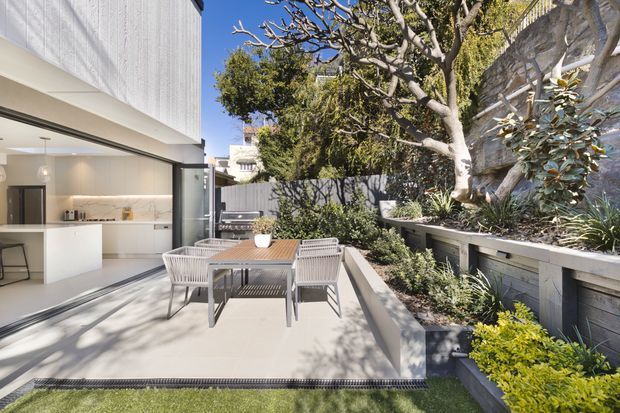 A bright contemporary outdoor dining terrace adjoining an open-plan white kitchen. The paved patio has a timber dining table with woven chairs, raised planted garden beds, a mature tree, and a textured masonry boundary wall. Large sliding glazing connects the terrace to the interior, while a narrow strip of lawn borders the foreground.