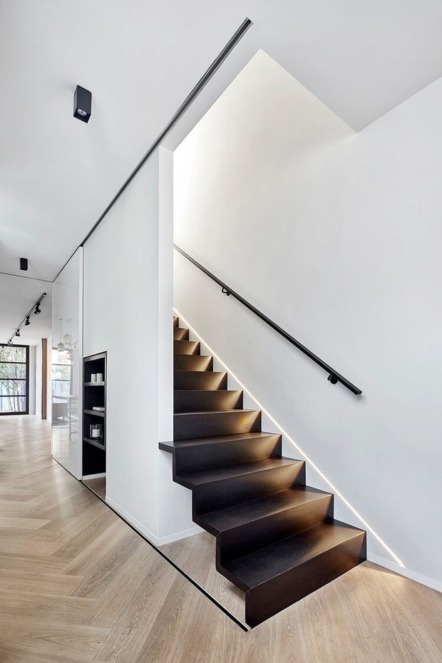 A minimalist contemporary interior focused on an open dark timber staircase with a slim black metal handrail and concealed LED lighting along the wall. The staircase rises beside a white partition wall and glass panels, with a light timber herringbone floor, recessed shelving, and track lighting extending into the adjacent hallway. The space is bright, clean, and sharply detailed with strong geometric lines.