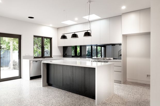 A bright contemporary kitchen with a large white waterfall-edge island, dark timber-look island cabinetry, white overhead and wall cabinetry, black-framed windows, stainless-steel appliances, and a terrazzo-style speckled floor. The room includes pendant lights, extensive glazing, and a glazed door opening to an outdoor area.