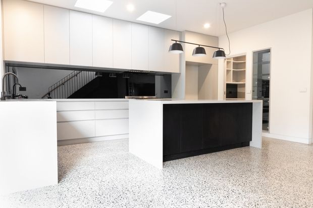 Bright contemporary kitchen with a large white island, dark contrasting island cabinetry, extensive white overhead and base joinery, integrated appliances, black pendant lights, and a polished terrazzo floor. A stairway is visible through the open area behind the kitchen.