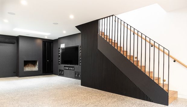 Contemporary media and living area beneath a staircase, featuring a dark built-in entertainment wall with fireplace and television, vertical black slatted screening, timber stair treads, and slim black metal balustrading. The room has a light carpet, white ceiling with recessed lighting, and a strong black-and-white contrast.