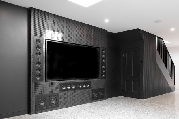 A monochrome contemporary home theatre/media room with a large wall-mounted television integrated into a perforated dark acoustic feature wall, surrounded by multiple built-in speakers. A dark paneled door and enclosed staircase with vertical balustrades are visible to the right. The room has a bright white ceiling with recessed lighting and a light speckled floor.