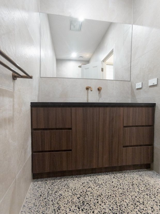 Bright contemporary bathroom vanity area with a wall-to-wall dark timber-look floating cabinet, black benchtop, brass wall-mounted tapware, large frameless mirror, pale stone-look wall tiles, and a terrazzo-style floor. A towel rail and double power outlets are visible.