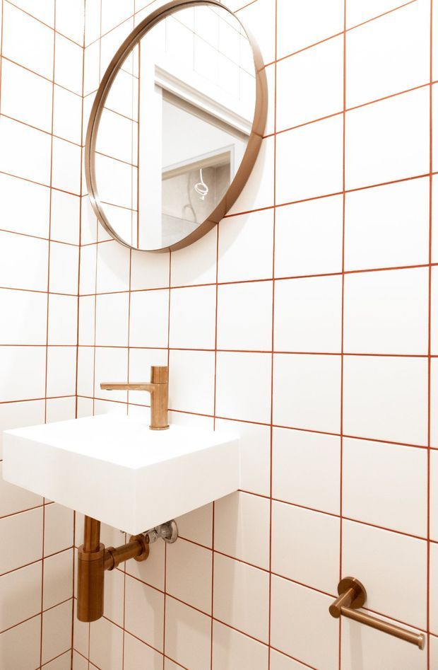 Compact bathroom vanity area with white square wall tiles outlined by terracotta-red grout, a white wall-mounted basin, exposed copper-toned plumbing, matching copper-toned minimalist tapware and towel rail, and a large round mirror with a warm metallic frame. The space is bright and graphic with a clean contemporary aesthetic.