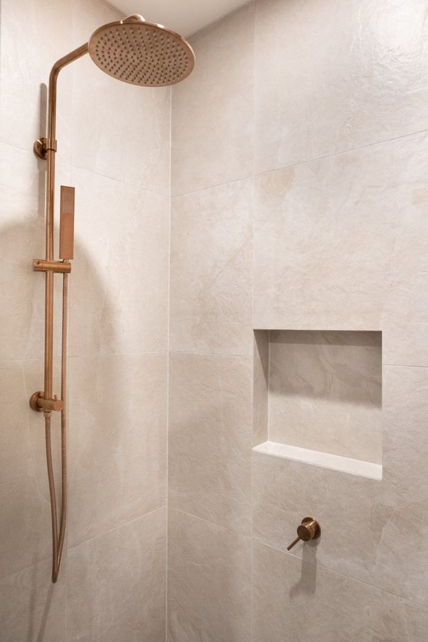 A close-up view of a minimalist walk-in shower enclosure with large pale beige stone-look wall tiles, a recessed wall niche, and a brushed bronze/copper-toned rain shower system with handheld shower and controls. The space is clean and uncluttered with a warm neutral palette.