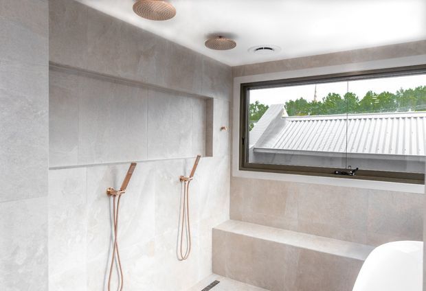 A bright contemporary bathroom with a large walk-in shower, dual wall-mounted copper-toned shower fittings, pale large-format tiled walls, a built-in tiled bench, and a wide framed window overlooking a corrugated metal roof and trees. The room has a clean, refined spa-like appearance.