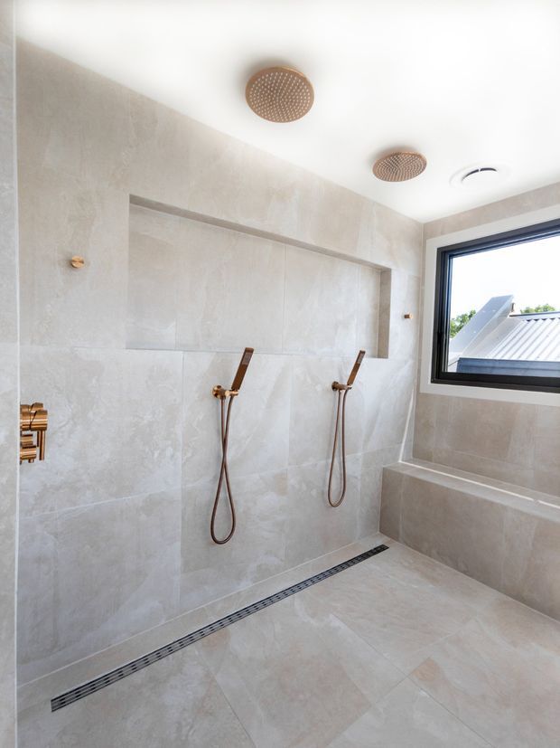 Bright contemporary bathroom featuring a large walk-in shower with dual handheld shower sets, two round ceiling-mounted rain showerheads, pale large-format stone-look wall and floor tiles, brushed brass fixtures, a linear floor drain, built-in bench, and a black-framed window.