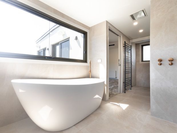 Bright contemporary bathroom featuring a freestanding white oval bathtub beneath a large black-framed window, pale stone-look wall and floor finishes, a walk-in shower area with a black heated towel rail, recessed ceiling lighting, and warm timber-toned wall hooks. An open doorway leads to a small adjacent area with patterned flooring.