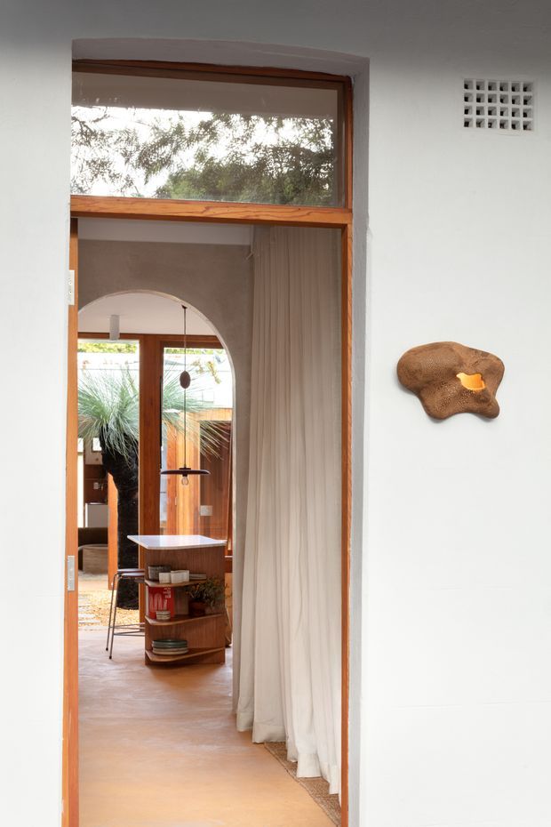 Interior view through a timber-framed glazed doorway into a warm, softly lit living or entry space. White plaster walls and a smooth earthy-toned floor frame an arched opening, timber doors, sheer curtains, a small built-in or freestanding timber shelving unit, and a sculptural wall artwork. A breeze-block ventilation panel adds a distinctive architectural detail, while vegetation is visible beyond the glazing.