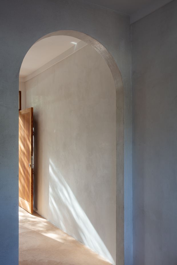 A softly lit interior passage framed by a large rounded archway. The space has pale textured plaster walls, a warm smooth floor, and a partially open timber door with vertical slats at the left. Sunlight creates diagonal highlights across the wall and floor, producing a calm, restrained architectural composition.