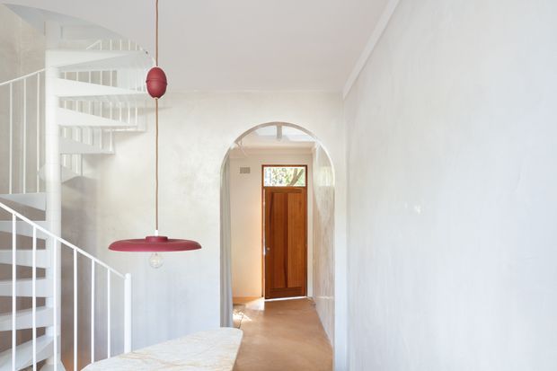 Bright minimalist residential hallway and entry area with white textured walls, a sculptural white spiral staircase and slim white balustrade on the left, and a warm timber front door framed by a rounded arch at the end of the passage. A red pendant light provides a vivid accent above a pale stone or terrazzo surface in the foreground.