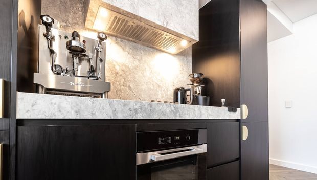 Close-up view of a contemporary kitchen coffee station and cooking area, featuring dark vertical-grain cabinetry, a pale marble-look benchtop and splashback, integrated oven, stainless-steel espresso machine, grinder, and a concealed rangehood. The space has clean-lined joinery and bright task lighting.