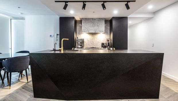 Contemporary open-plan kitchen dominated by a large dark stone island with integrated seating, a brass-toned faucet, dark full-height cabinetry and a textured grey stone backsplash. Track lighting and recessed ceiling lights illuminate the space, with a dining area visible to the left and light timber flooring throughout.