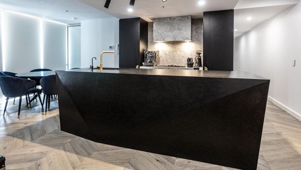 Contemporary open-plan kitchen dominated by a large sculptural black island with a faceted waterfall-style front. The kitchen includes dark cabinetry, a textured grey stone backsplash, integrated lighting, a brass-toned tap, and light herringbone timber flooring. A small dining area with dark chairs is visible behind the island.