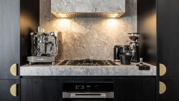 Close-up view of a compact, highly finished kitchen coffee and cooking station. Dark timber or black cabinetry with large rounded brass-toned pulls frames a veined grey stone benchtop and matching stone splashback. A stainless steel espresso machine and grinder sit beside a black gas cooktop, integrated oven, kettle, and coffee accessories. Warm under-cabinet lighting highlights the stone surface.