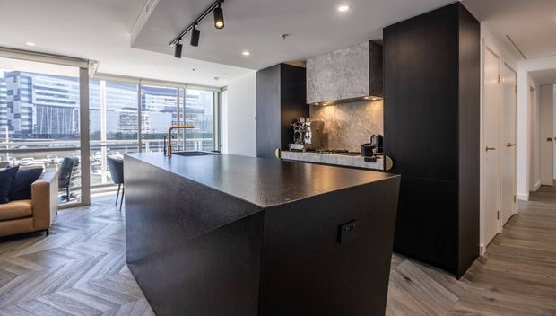 Contemporary open-plan kitchen featuring a large dark island with a textured stone benchtop, integrated dark cabinetry, a stone splashback, timber herringbone flooring, and track lighting. Floor-to-ceiling glazing opens toward a city view and adjacent living/dining area, with a hallway extending to the right.
