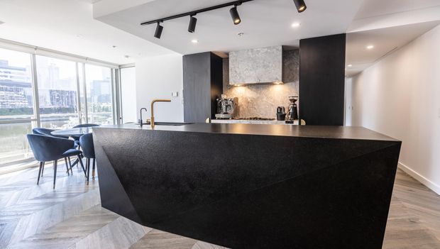 Contemporary open-plan kitchen dominated by a large angular black island with a dark stone-look benchtop and waterfall sides. The kitchen features dark vertical cabinetry, a textured stone splashback, integrated track lighting, timber-toned herringbone flooring, and a dining area beside full-height corner glazing.