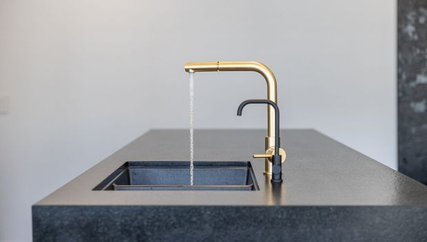 Close-up of a contemporary kitchen island with a dark stone benchtop, integrated rectangular sink, and two contrasting mixer taps: a brushed brass gooseneck tap running water and a smaller dark tap beside it. The background is minimal with neutral grey walls and a dark stone panel.