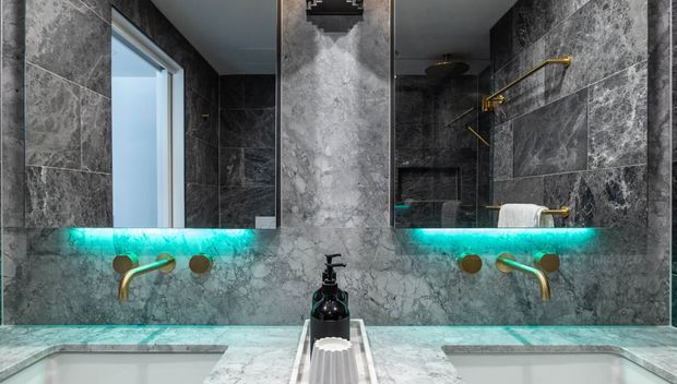 Luxurious contemporary bathroom vanity with twin rectangular basins, a large mirror, grey marble-look wall and benchtop surfaces, and distinctive brushed gold wall-mounted taps and fittings. Turquoise LED lighting glows beneath the mirror, creating a dramatic contrast against the dark stone finish.