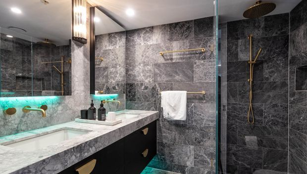 Luxurious contemporary bathroom featuring dark marble-look wall and floor tiles, a double vanity with black cabinetry and a pale stone benchtop, large mirrored cabinetry, glass-enclosed shower, and brushed gold fixtures. Dramatic teal under-vanity lighting adds a vivid accent.