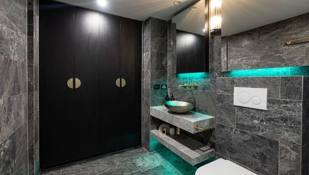 A dramatic contemporary bathroom clad in dark grey marble-look tiles, with black double doors, a floating stone vanity, vessel basin, wall-mounted toilet, illuminated turquoise accent lighting, and brass hardware.
