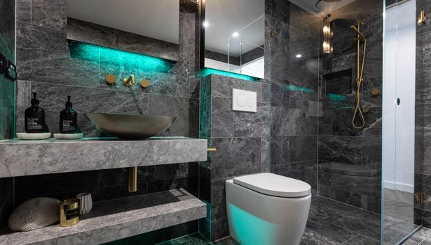 A dramatic contemporary bathroom with dark grey marble-look wall and floor surfaces, a floating stone vanity with open shelving, vessel basin, wall-mounted tapware, concealed cistern toilet, and glass-enclosed shower. Integrated turquoise LED lighting creates a vivid accent against the dark finishes, with brushed brass shower and fittings adding warmth.