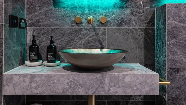 Close-up of a luxurious bathroom vanity featuring a vessel basin on a thick stone countertop, dark marble-look wall tiles, black fixtures and accessories, and atmospheric turquoise accent lighting.