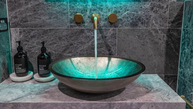Close-up view of a bathroom vanity featuring a freestanding vessel basin beneath a wall-mounted brass-toned faucet. Dark grey marble-look wall and counter surfaces, matching dark tile side walls, and teal lighting/reflections create a dramatic, luxurious atmosphere. Soap dispensers sit beside the basin.