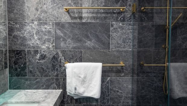 Close-up of a dark, luxurious bathroom shower area clad in varied grey veined stone tiles. Brass or antique-gold towel rails, shower fixtures and a handheld shower create warm metallic accents, while a white towel hangs in the center. A pale stone ledge or bench is visible at lower left, and reflective glass appears at right.