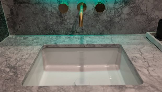 Close-up of a luxurious bathroom vanity featuring a rectangular integrated white basin set into a grey veined marble countertop and matching marble splashback. A wall-mounted brushed brass/gold tap and fittings provide a warm metallic accent, with subtle teal ambient lighting.
