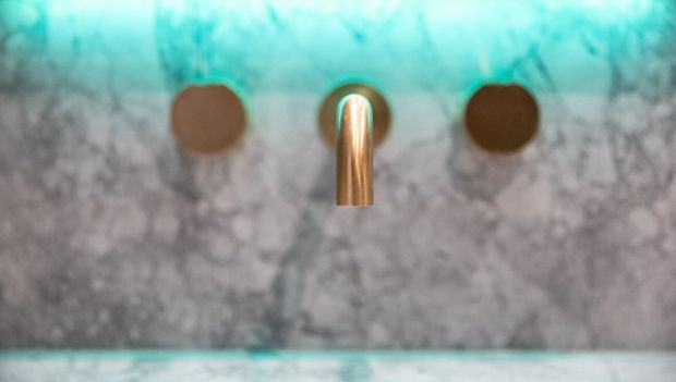 Close-up of a wall-mounted bathroom tap set with a central brushed brass spout and two round brass controls, set against a grey veined stone wall and beneath a turquoise-tinted stone or glass surface.