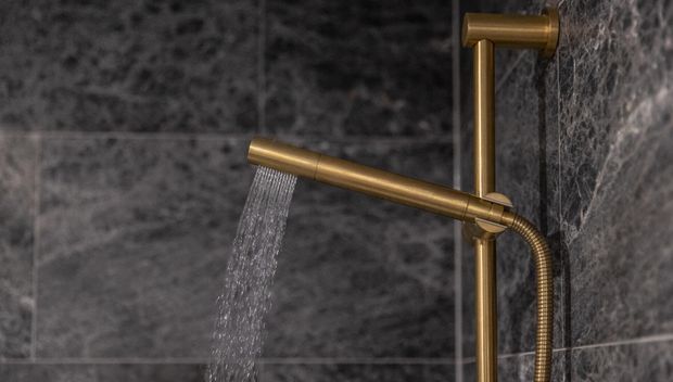 Close-up of a contemporary shower system mounted against dark grey stone-look tiled walls. The fixture has a warm brushed brass finish, with a horizontal handheld shower rail, overhead shower outlet, and visible flowing water.