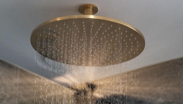 Close-up of a large round rainfall showerhead mounted to the ceiling, with water streaming from numerous nozzles. The showerhead has a warm brushed brass or gold-toned finish, set against a pale ceiling and tiled or stone bathroom surfaces.