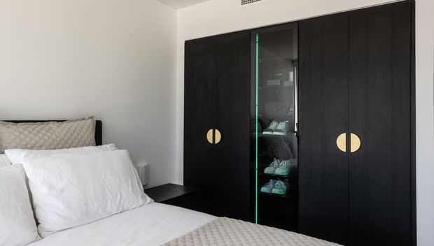 A contemporary bedroom with a neatly made bed in white bedding and a quilted neutral throw. A large wall-to-wall dark timber wardrobe dominates the room, featuring integrated circular brass-toned handles and a central glass-front display section. The room has white walls, dark bedside furniture, and a clean, restrained palette.