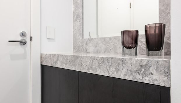 Close-up of a refined bathroom vanity with dark timber cabinetry, a thick grey-white veined stone benchtop and matching stone mirror surround. Two smoked glass vessels sit on the counter, with a white door and minimal hardware visible at left.