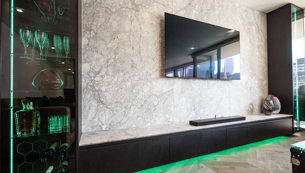 Contemporary media wall with a wall-mounted television, long dark built-in cabinetry, and a dramatic grey veined stone feature wall. Illuminated glass display cabinets with green LED lighting flank the arrangement, while a chevron timber floor adds warmth. The space has a refined, high-contrast entertainment-room aesthetic.