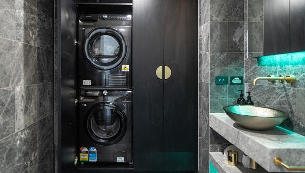 Compact, moody bathroom laundry zone featuring stacked black front-loading washer and dryer in dark built-in cabinetry, beside a vessel basin on a stone vanity. Grey stone-look tiled walls, dark joinery, brass hardware, and turquoise under-cabinet lighting create a dramatic contemporary fit-out.