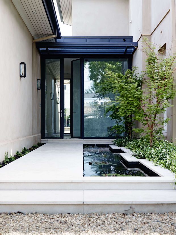 A refined residential entry courtyard viewed from outside, with broad pale concrete steps and paving leading to dark-framed glazed entry doors. A shallow rectangular water feature with aquatic plants runs alongside the walkway, bordered by clipped greenery and slender trees. The courtyard is enclosed by pale rendered walls and features a dark blue-black steel-and-glass canopy, creating a calm, modern architectural composition.