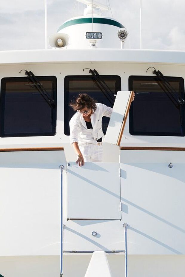 A bright exterior view of a white motor yacht, focused on the raised aft or side boarding entrance. The vessel has dark rectangular windows, polished metal handrails, timber trim, marine equipment on the upper deck, and a person stepping through the hatch. The image is dominated by the boat's exterior rather than an architectural interior.