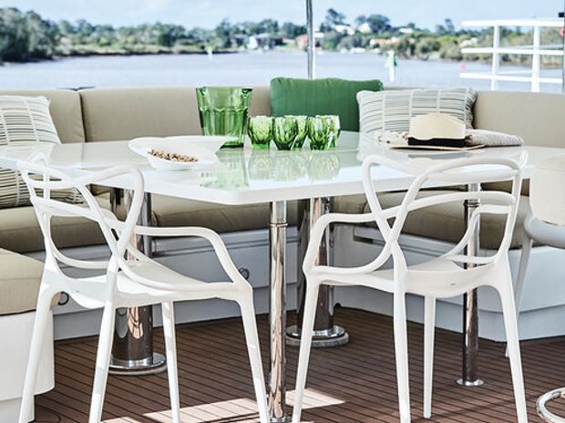 Bright contemporary dining area aboard a boat or waterfront vessel, featuring a white round table with white sculptural chairs, a pale upholstered banquette, green glassware, chrome pedestal supports, and dark timber-style decking. Water and shoreline are visible through the open glazing.