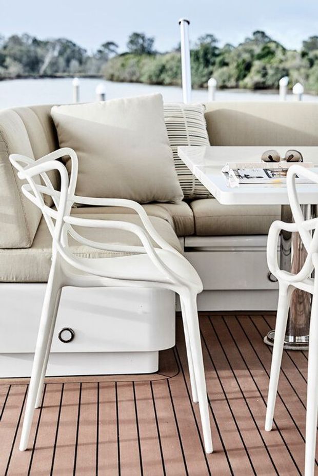 A bright, refined outdoor boat deck or waterside seating area with a cream upholstered built-in bench, white sculptural dining chairs, a compact white table, neutral cushions, and warm timber-look slatted decking. Calm water and greenery are visible beyond the low railing.