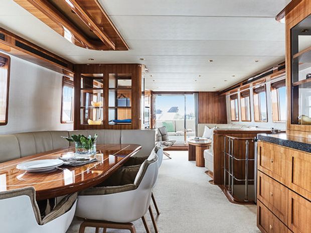 A bright, refined open-plan yacht interior combining a dining area, lounge and compact galley kitchen. Rich polished timber cabinetry and wall panelling contrast with light upholstered seating, a pale carpeted floor and a white paneled ceiling. Large windows provide abundant daylight and views outside, while built-in joinery and curved furniture emphasize the marine setting.