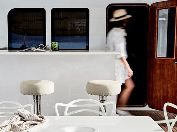 Bright contemporary kitchen and dining area with a glossy white island or breakfast counter, two upholstered bar stools, white sculptural dining chairs, and a white dining table in the foreground. Dark rounded-rectangle windows, a dark timber entry door, and a blurred figure add contrast to the predominantly white interior.