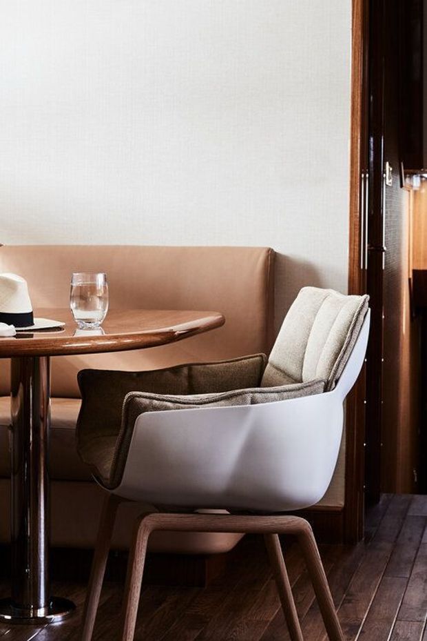 A close-up interior view of a refined dining nook featuring a curved upholstered banquette, a white molded armchair with timber legs, and a small round wood-topped table with a chrome pedestal. Dark timber wall framing and dark timber flooring create a warm, sophisticated setting, with neutral upholstery and subtle tableware.
